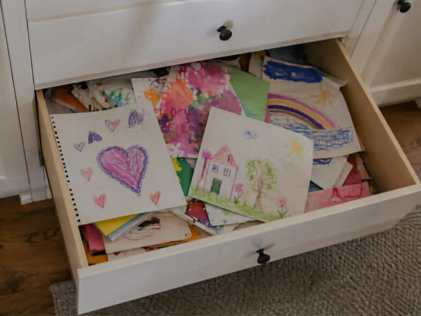 Open dresser drawer overflowing with stacks of children's drawings and paintings